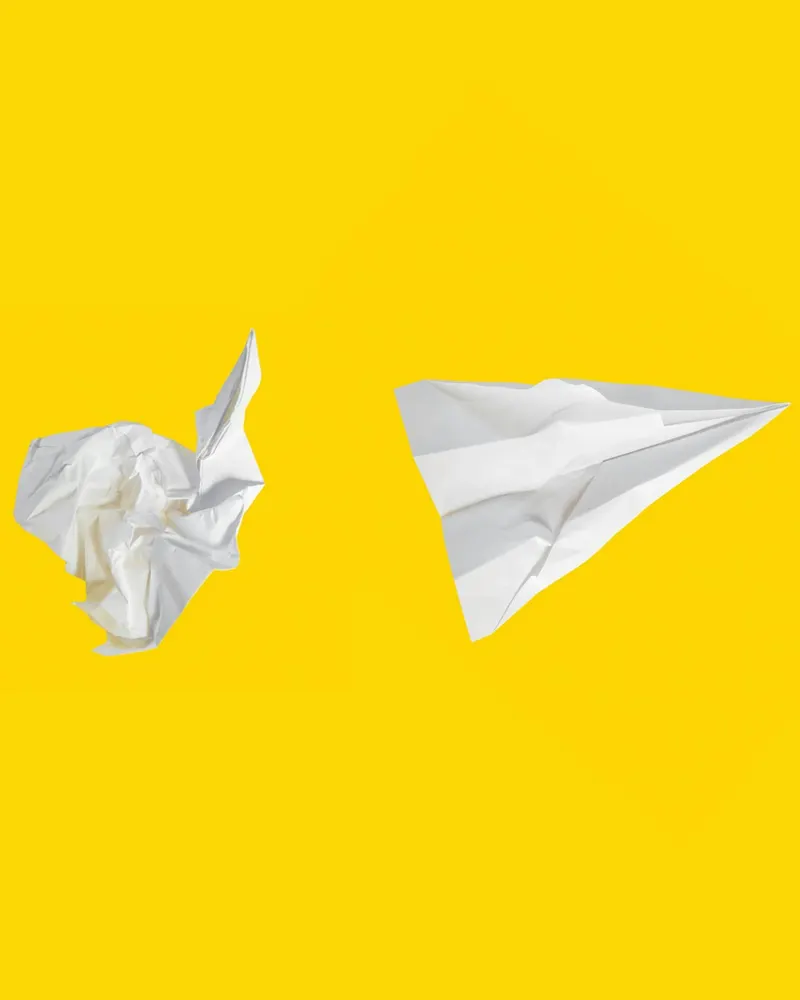 A crumpled ball of white paper next to a folded paper plane on a bright yellow ground