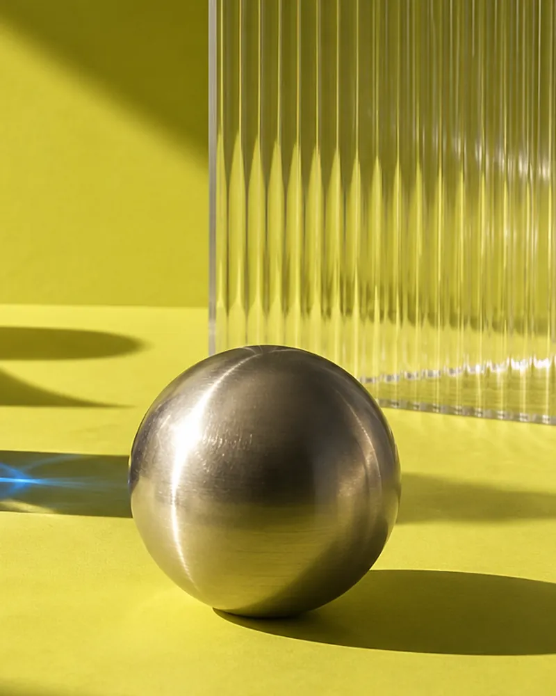 A brushed metal sphere in front of a fluted glass panel on a yellow-green surface, with hard shadows