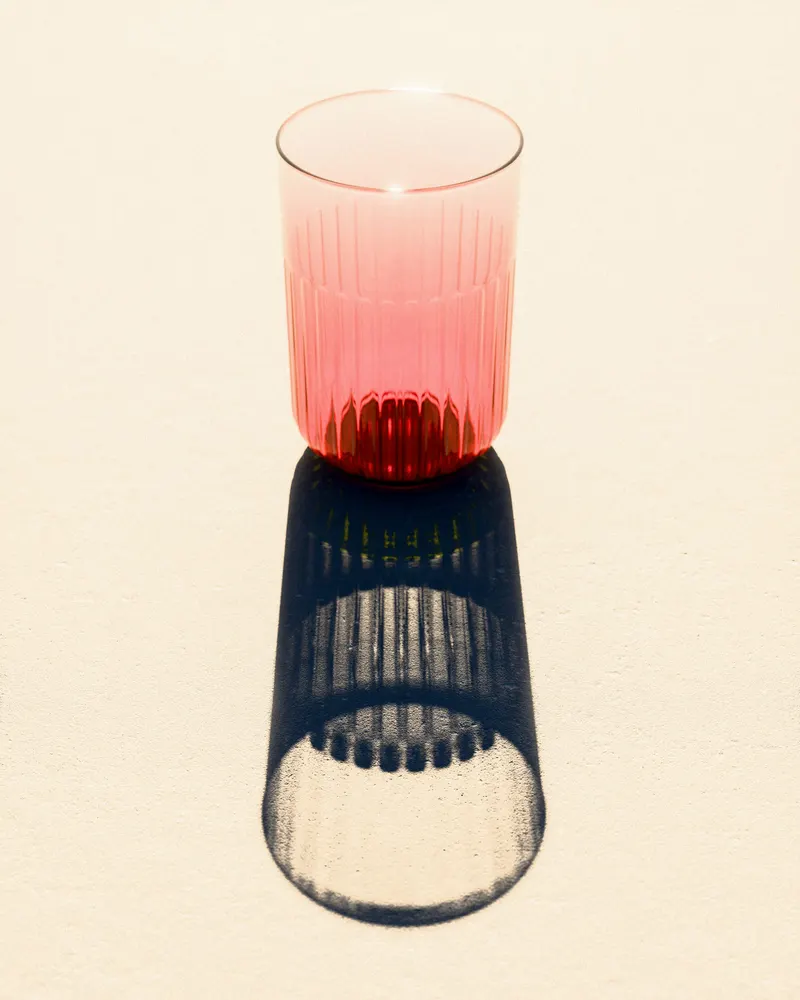 A fluted glass, pink at the rim, casting a long shadow on a cream surface under hard light