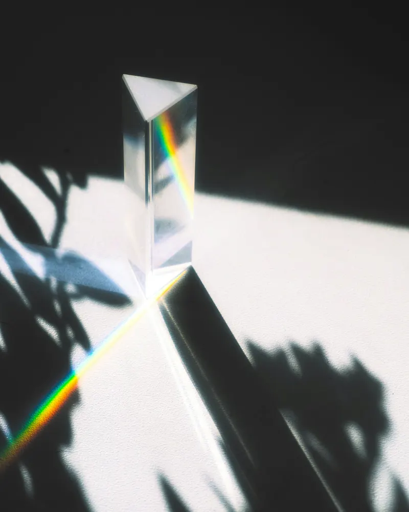 A glass prism on a white surface splitting sunlight into a thin rainbow, with leaf shadows around it